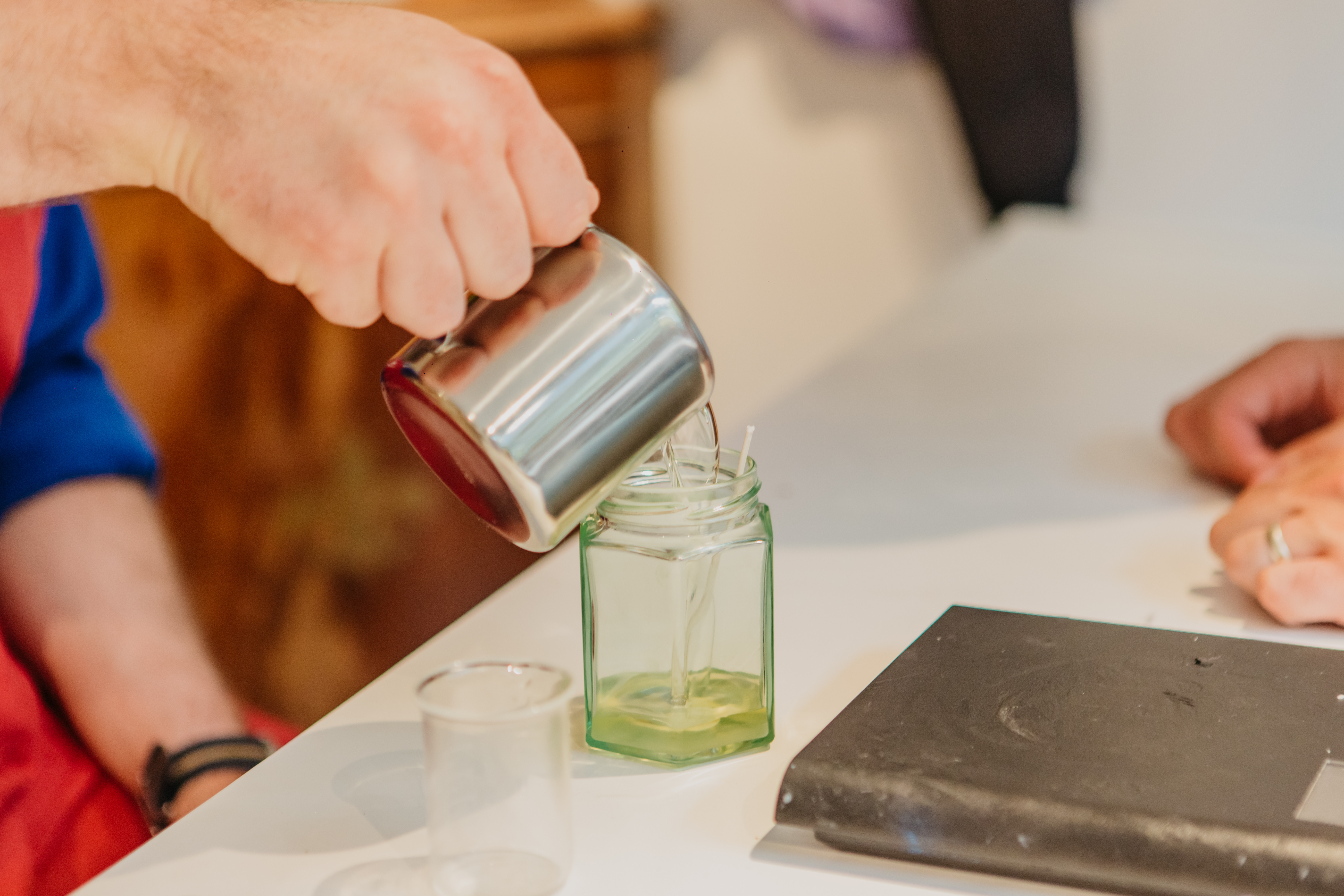 A man pours soy wax into a glass candle jar from a small metal jug